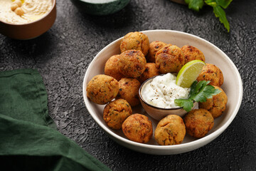 Traditional oriental chickpea deep fried falafel in a deep bowl with tzatziki yoghurt sauce, hummus, fresh lime and green cilantro on black surface