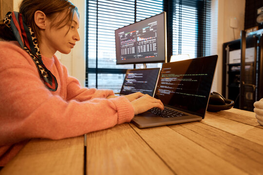 Woman writing programming code on laptops and desktop computer at cozy home workplace. Concept of remote programmer work at home