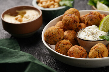 Traditional oriental chickpea deep fried falafel in a deep bowl with tzatziki yoghurt sauce, hummus, fresh lime and green cilantro on black surface
