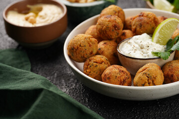 Traditional oriental chickpea deep fried falafel in a deep bowl with tzatziki yoghurt sauce, hummus, fresh lime and green cilantro on black surface