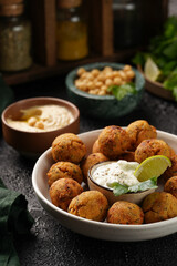 Traditional oriental chickpea deep fried falafel in a deep bowl with tzatziki yoghurt sauce, hummus, fresh lime and green cilantro on black surface