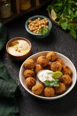 Traditional oriental chickpea deep fried falafel in a deep bowl with tzatziki yoghurt sauce, hummus, fresh lime and green cilantro on black surface