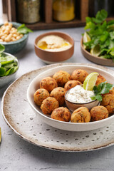 Traditional oriental chickpea deep fried falafel in a deep bowl with tzatziki yoghurt sauce, hummus, fresh lime and green cilantro on light grey concrete surface