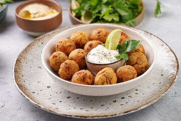 Traditional oriental chickpea deep fried falafel in a deep bowl with tzatziki yoghurt sauce, hummus, fresh lime and green cilantro on light grey concrete surface
