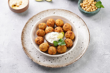 Traditional oriental chickpea deep fried falafel in a deep bowl with tzatziki yoghurt sauce, hummus, fresh lime and green cilantro on light grey concrete surface