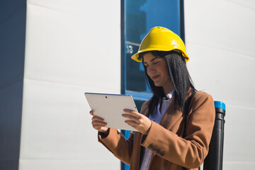 Young beautiful woman in brown coat, yellow workman's helmet and blueprint tube, working with her tablet. Business concept, architect, construction, working woman, empowerment.