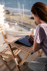 A young woman in headphones is working on tablet on the balcony near the sea