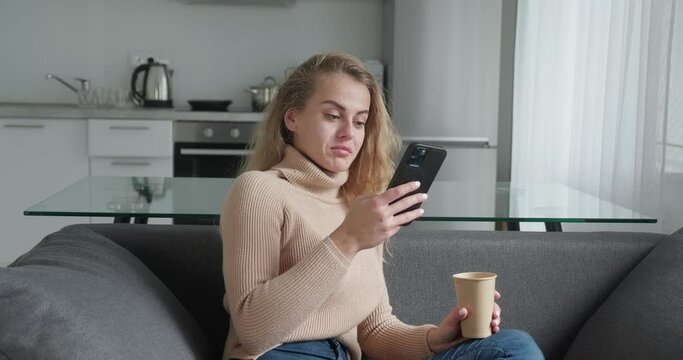Young blonde woman at home, smiling girl using app and browsing on the internet, typing message on smartphone sitting on cozy sofa, drinking cofee, beautiful lady having pleasant funny conversation