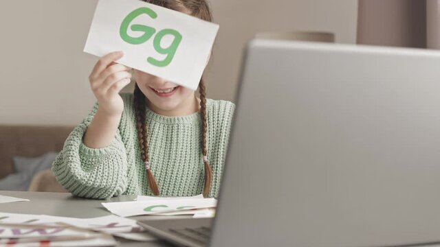 Waist up slowmo shot of little girl with printed letter cards having online lesson of speech therapy via laptop from home