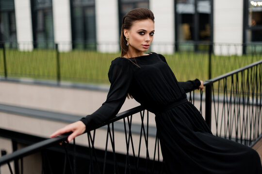 Gorgeous woman in stylish black dress posing for the photo, looking directly at the camera, showing self confidence. Beautiful female working as fashion model, having a photoshoot for magazine cover