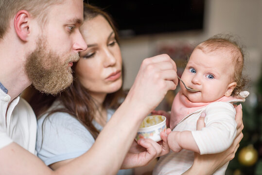 Father and mother feed their baby fruit puree in the kitchen from a spoon