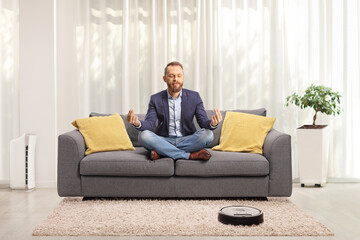 Young man meditating on a sofa and a robot vacuum cleaning the carpet