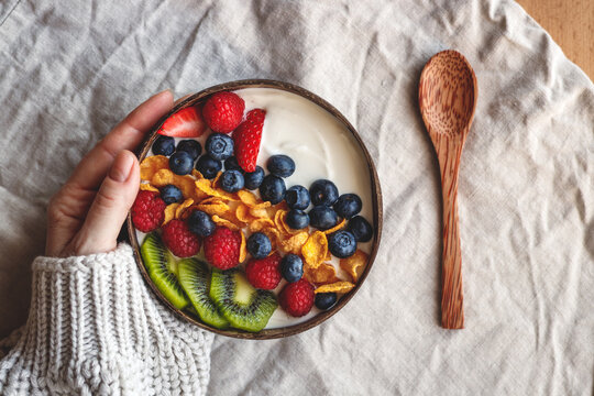 Healthy breakfast in coconut bowl on table with linen tablecloth. Yogurt with corn flakes and blueberry, strawberry, kiwi and raspberry. Womans hand wearing sweater. Vegetarian food