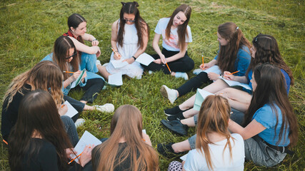 A group of female students are sitting in a circle on a meadow for collective work with notebooks.