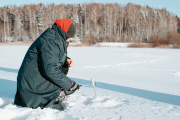 Fototapeta na wymiar Winter fishing, active leisure, hobby and sport concept. Side view of man fisherman catching fish, sitting on frozen water holding fishing rod against of snowy forest on sunny day. Copy space