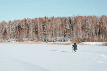 Man fisherman drilling hole in frozen water on ice while standing against background of snowy forest in nature, copy space. Winter fishing, active leisure, hobbies and recreation concept