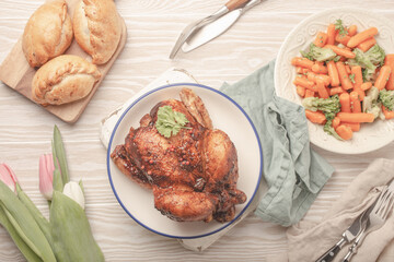 Easter food on white rustic table: pastel colored eggs, roasted chicken and vegetables, buns and spring flowers tulips top view flay lay, Easter family dinner meal with festive dishes 