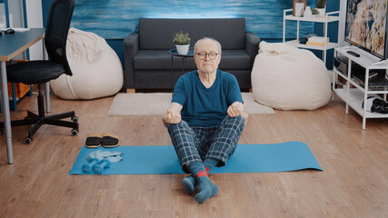 Fototapeta premium Old person sitting in lotus position with closed eyes on mat to practice calm meditation. Senior man using yoga pose to meditate and find relaxing balance. Pensioner doing wellness activity