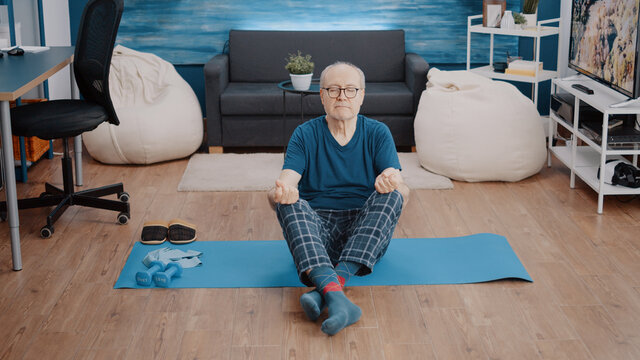Old person sitting in lotus position with closed eyes on mat to practice calm meditation. Senior man using yoga pose to meditate and find relaxing balance. Pensioner doing wellness activity