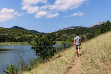 A young man is walking alone by the lake with a mountain landscape