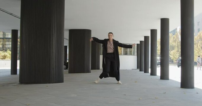 Attractive businesswoman in formal black trench is dancing outside the office, financial district, modern constructivist building with columns on background. The concept of urban city scene