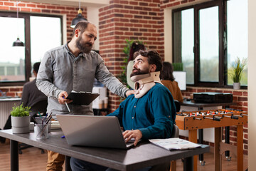 Millennial freelancer recovering after accident having neck pain wearing medical neck collar while working in brick wall startup office. Businessmen analyzing company turnover discussing business