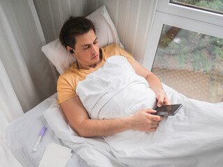 Ill man is lying in bed with thermometer. Man is searching online for disease symptoms or texting to his doctor. using wireless technology in case of temporary disability.