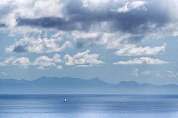 a small white sailing ship sails in the vast ocean. In the background you can see an island in a day with a sky with many clouds. Concept of tranquility, smallness, immensity, greatness, loneliness