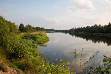 Autumn forest behind the lake. Sky with sun and white clouds. Red-green forest.