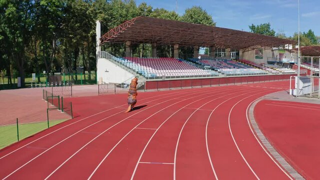 Following the person in funny dinosaur costume running along the tack. Tirex dinosaur jogging around the stadium.