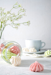 Meringue cookies are white, pink and light green in a transparent jar on a light table. A white cup with coffee in the background. White flowers in a transparent vase on the table