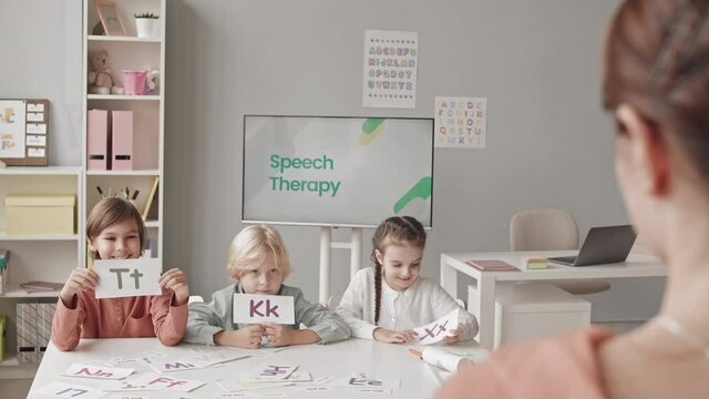 Medium slowmo shot of elementary school kids showing cards with colorful letters to female speech therapist during lesson in classroom