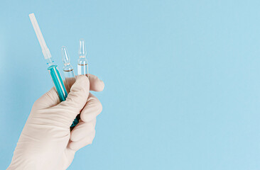 Hand in protective glove holding ampules with vaccine and syringe on blue background. Copy space
