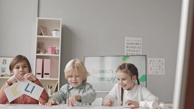 Waist up slowmo shot of three cheerful elementary kids learning alphabet having speech therapy lesson in classroom