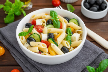 Healthy pasta salad with tomatoes, cucumbers, red onions, olives and cheese in a bowl on a dark wooden background. Copy space.