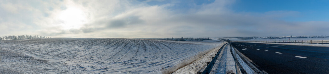 Fototapeta premium Country road through winter rural scene. Black clean asphalt. Winter road going through snow-covered fields.