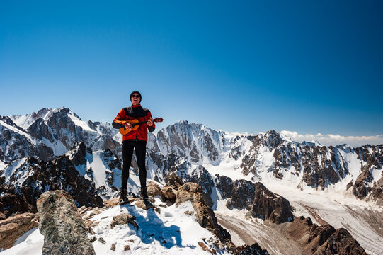 Young man trekker playing guitar and singing song on top of Pik Uchitel peak . Ala Archa Alpine National Park Landscape near Bishkek, Tian Shan Mountain Range, Kyrgyzstan, Central Asia.