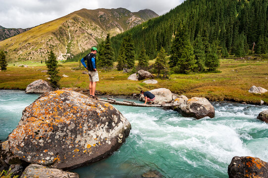 Young man and woman crossing wild turquoise river on tiny wooden bridge on trail to Ala Kul lake. Karakol valley, Issyk-kul region, Ala-kul lake Terskey Alatau mountain range, Kyrgyzstan, Central Asia