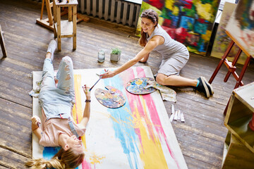 Funny female mastery with canvas and palette for mixing acrylic colors enjoying time for creating pictures resting in art studio and smiling while communicate about hobby, cheerful friends