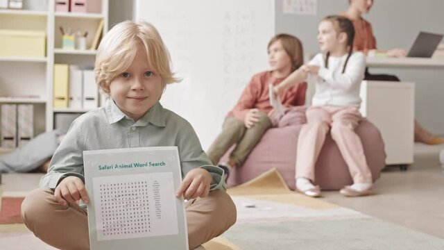 Slowmo portrait of 7-year-old Caucasian boy posing for camera with word search worksheet in hands having lesson of speech therapy with other kids and female teacher