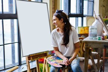 Happy Caucaisan woman with palette and paintbrush laughing during leisure pastime for creating sketch, joyful hipster girl with equipment for drawing enjoying own hobby during weekend in art studio