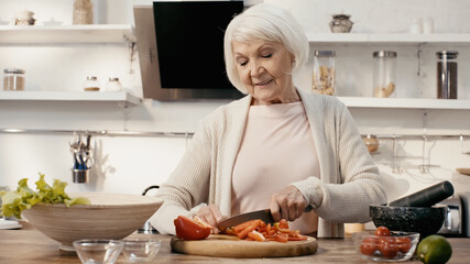 joyful senior woman cutting bell pepper near fresh cherry tomatoes and lime in kitchen.