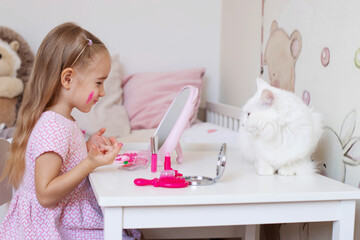 A little girl sits at a children's table in front of a mirror and paints her lips.