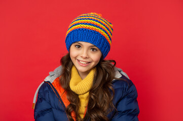 cheerful teen girl in knitted winter hat and down coat on red background, teenager