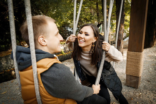 Cheerful young mixed race couple looking at each other while sitting on a swing outdoor. - Powered by Adobe