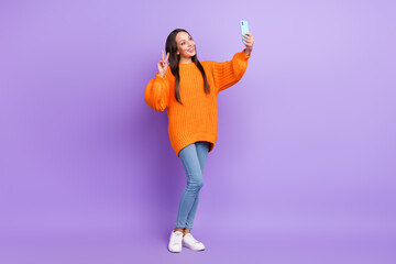 Fototapeta na wymiar Photo of friendly cute lady hold phone make selfie show v-sign wear orange sweater isolated purple color background