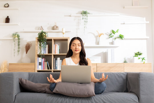 Clam Of Asian Young Woman Doing Yoga Lotus Pose To Meditation And Relax On Couch During Work Online At Home.Happiness Female Break After Worked Close Her Eyes And Deep Breath With Yoga So Peaceful.