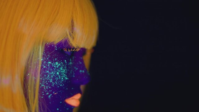 Young woman with bright glowing neon makeup wearing orange wig dancing over black studio background, empty space