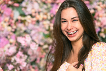 Young beautiful woman portrait. Female, girl smiles, smile with braces on a pink background with flowers. Copy space for billboard, Beauty and freshness of spring