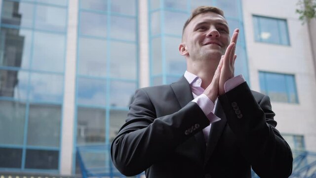 Portrait of excited smiling businessman gesturing victory standing at business center outdoors. Positive handsome Caucasian man in elegant suit smiling looking away. Good deal concept